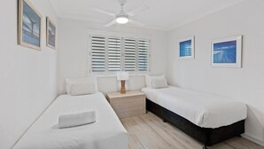 Un dormitorio con dos camas individuales, ventilador de techo, ventana con contraventanas y cuadros en las paredes.