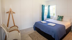 A bedroom with a bed, a chair, a hanging light fixture, and a window with curtains. - Cabana - One of the only original Straddie beach shacks left standing. (Point Lookout)