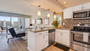 A modern kitchen with white cabinets, a stainless steel oven, and a microwave. There is a dining area with a black leather couch and a view of the city through large windows. - Ilikai 1128 Ocean/Lagoon Views / Free Parking / King Bed, Q Sofa Bed (Waikiki)