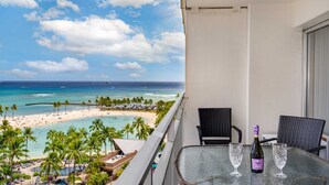 A balcony with a table, two chairs, and a bottle of wine. The view includes a beach, palm trees, and the ocean. - Ilikai 1128 Ocean/Lagoon Views / Free Parking / King Bed, Q Sofa Bed (Waikiki)