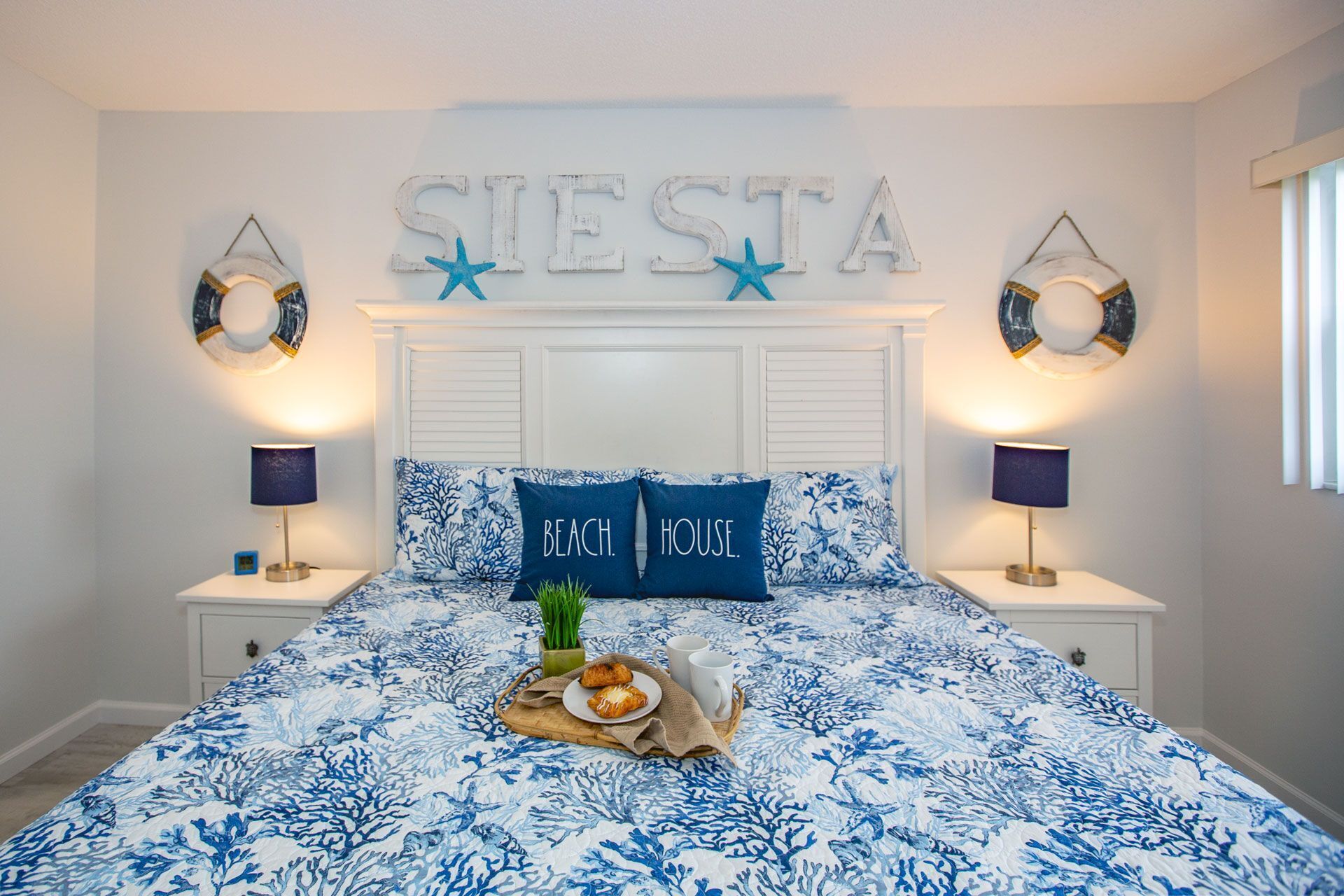 A bedroom with a bed, two nightstands, a wall-mounted sign reading "SIESTA" with star decorations, and a breakfast tray on the bed.