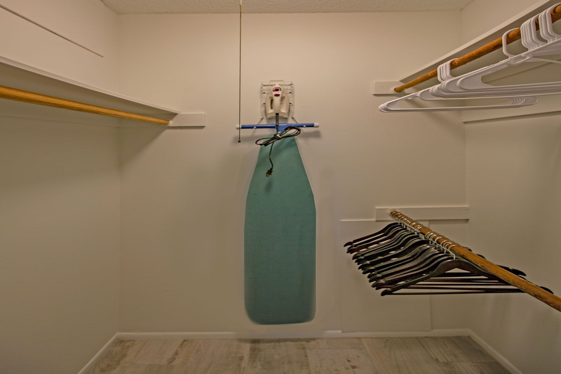 A small, narrow closet with a hanging rack for clothes, a blue towel, and a white wall with a wooden floor.