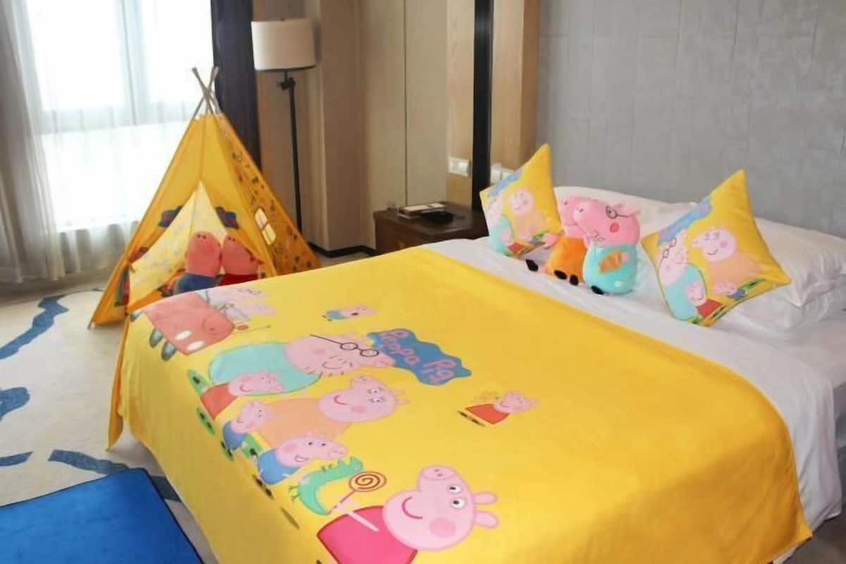 Une chambre d’hôtel avec un lit recouvert d’une couverture jaune ornée de personnages de dessins animés, un couvre-lit blanc et plusieurs oreillers aux motifs similaires.