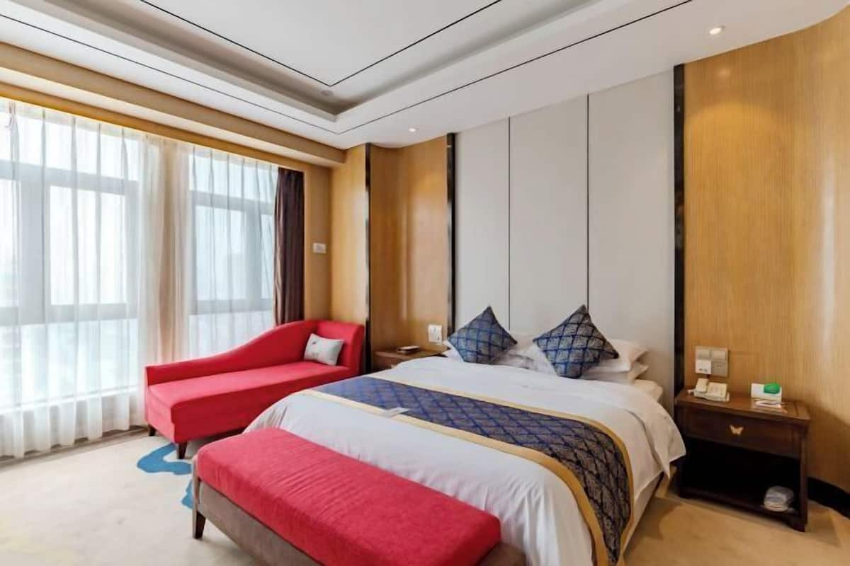 Une chambre d’hôtel avec un grand lit, un fauteuil rouge, une table de chevet avec une lampe et une grande fenêtre ornée de rideaux légers.