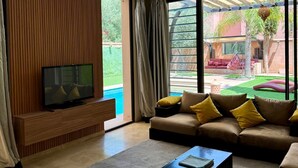 Una sala de estar moderna con televisor, sofá, mesa de centro, ventanas amplias y vistas a una piscina y zona exterior.