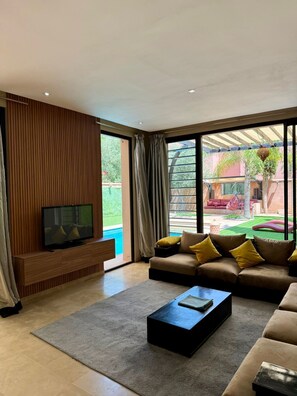 A modern living room with a TV, sofa, and coffee table, large windows, and a view of a pool and outdoor area. - Best 2Bd Room Villa in Al Maaden (Marrakesh-Safi)
