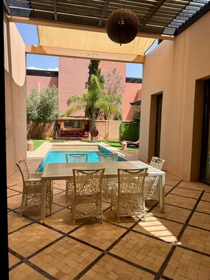 A covered patio with a pool, wicker furniture, and a view of a building with a red facade. - Best 2Bd Room Villa in Al Maaden (Marrakesh-Safi)
