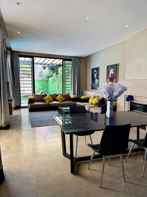 A modern living room with a sofa, a dining table, and a large window. - Best 2Bd Room Villa in Al Maaden (Marrakesh-Safi)