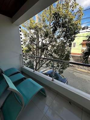 A balcony with green chairs, a railing, and a view of a tree and buildings.