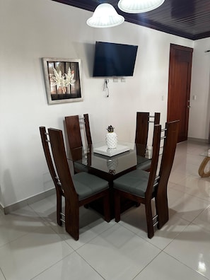 Een eetgedeelte met een tafel met glazen blad, houten stoelen, een ingelijste afbeelding en een aan de muur bevestigde televisie.