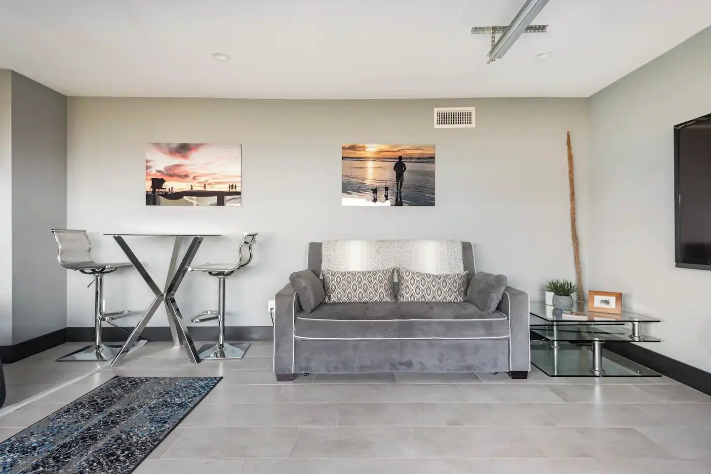 Un salon moderne avec un canapé gris, une table basse en verre, une chaise en métal et deux tableaux accrochés au mur.