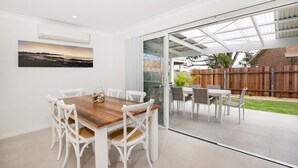 Un comedor con una mesa de madera y sillas blancas, un vidrio que da al exterior y vista a un patio trasero con valla de madera y follaje.