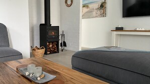 Una sala de estar acogedora con chimenea, sofás grises, una mesa de madera con bebidas y un cuadro con motivos playeros en la pared.