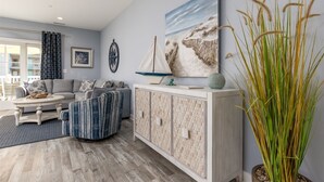 A modern living room with a grey sofa, a patterned ottoman, a white cabinet with a woven design, and a large painting of a beach scene.
