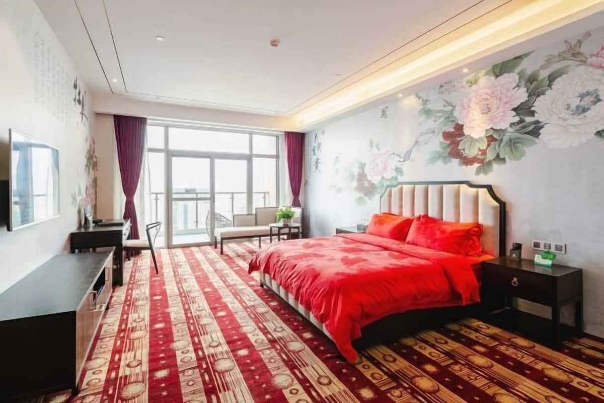 A bedroom with a bed, a desk, a chair, a nightstand, and a large window.