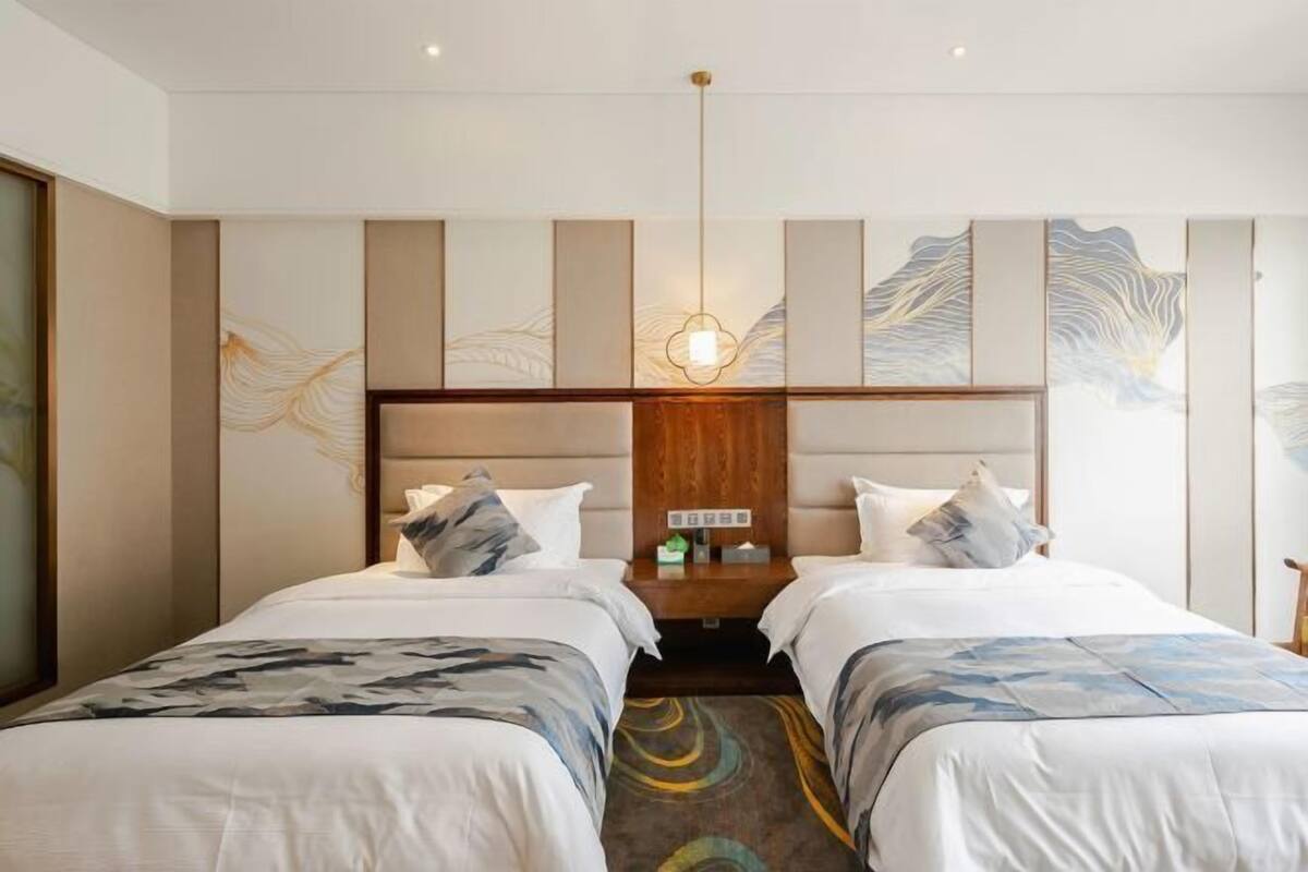 A hotel room with two beds, a wooden headboard, a wall art design, and a central light fixture.