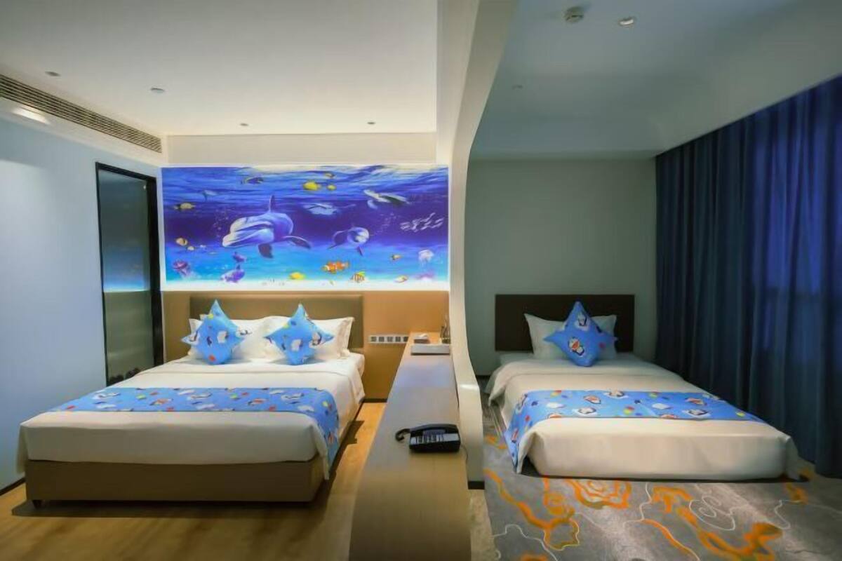 A hotel room with two beds, a large mural of an underwater scene, and a view of the ocean through the window.