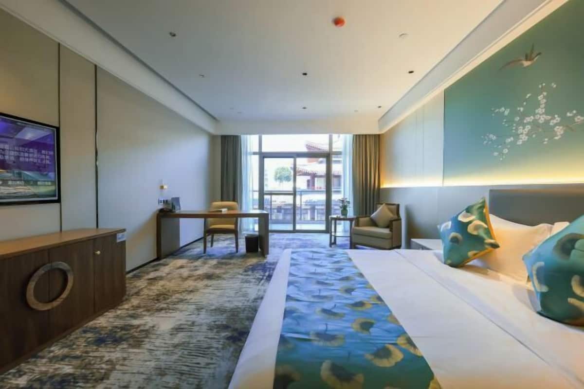 A hotel room with a bed, a desk, a chair, a TV, and a view of the outside.