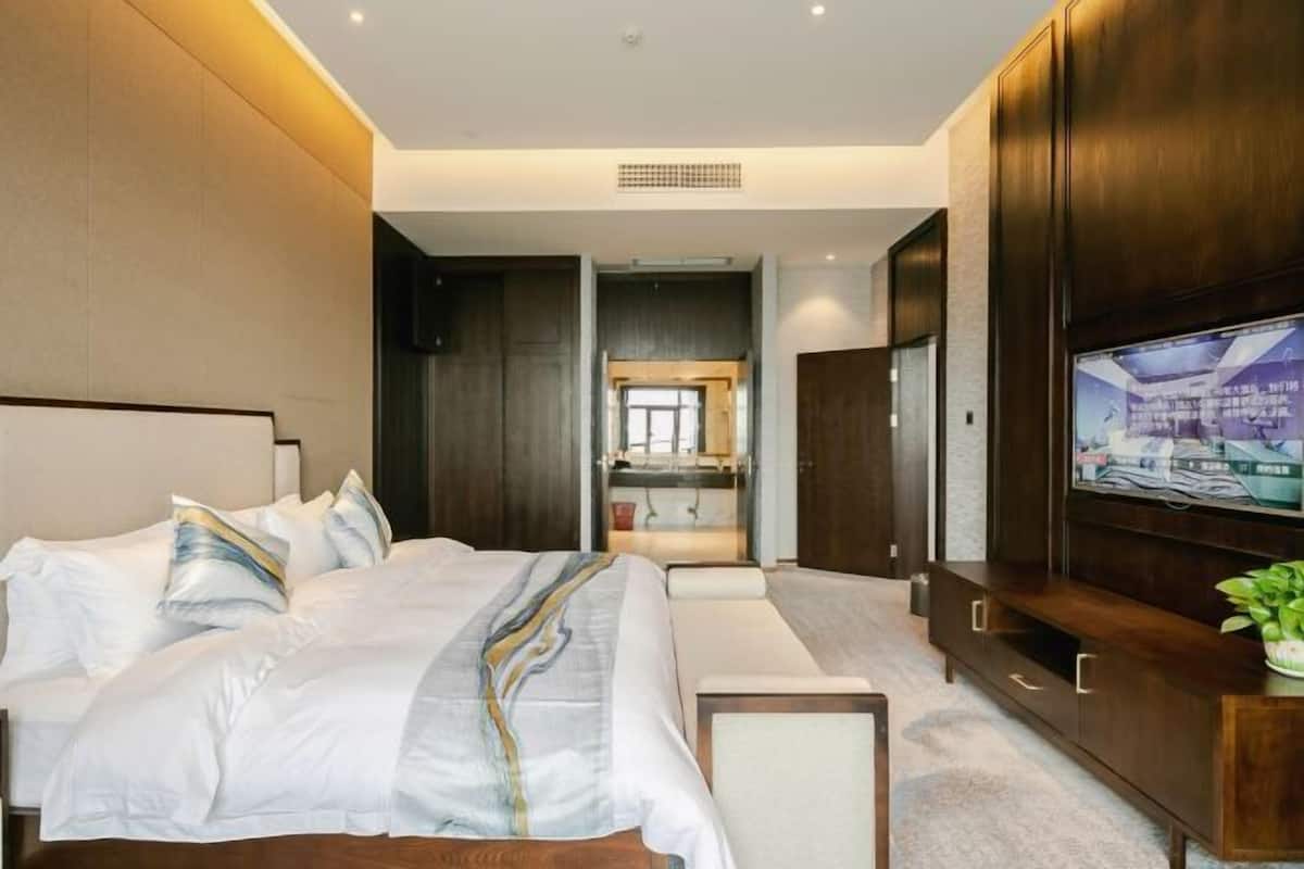 A modern hotel room with a large bed, a television, a wooden cabinet, and a view of the kitchen.