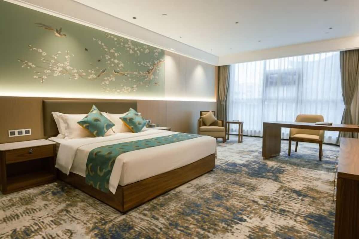 A hotel room with a large bed, a desk, a chair, and a view of the city through the window.