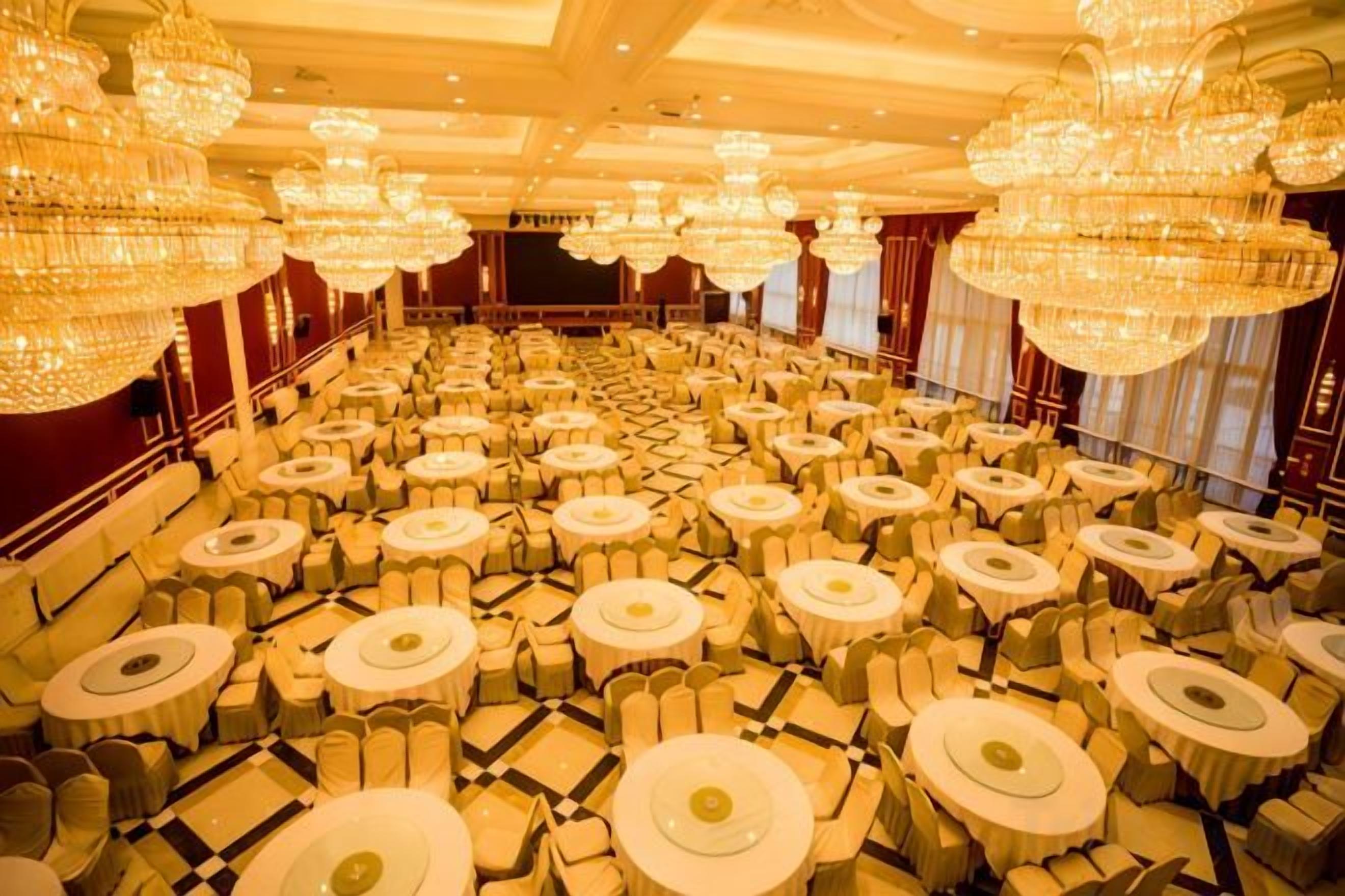 Un salón de banquetes con filas de mesas redondas y sillas, grandes candelabros y un escenario al frente.