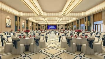 A large banquet hall with rows of tables and chairs, a stage at the front, and a large screen.