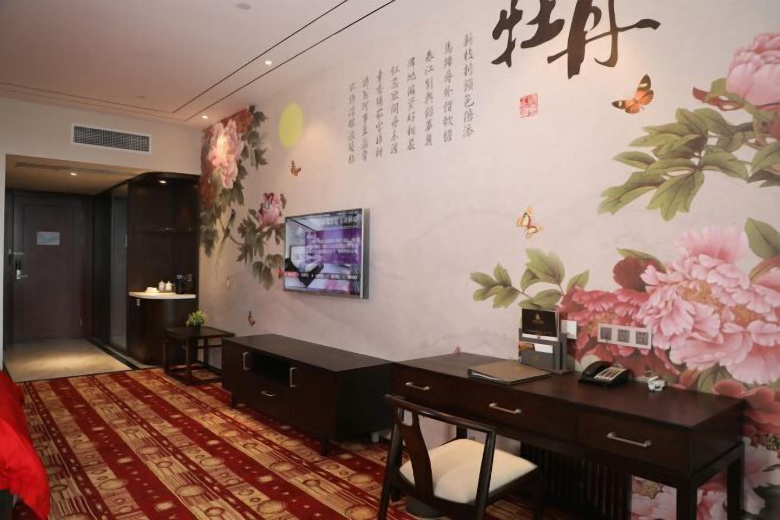 A hotel room with a TV, a desk, a chair, a bed, and a wall with floral and calligraphy decorations.