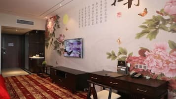A hotel room with a TV, a desk, a chair, a bed, and a wall with floral and calligraphy decorations.
