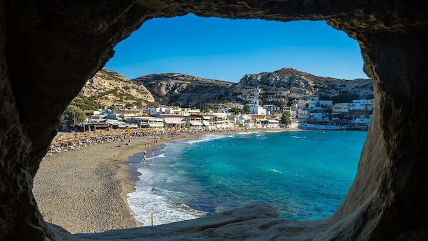 A coastal town viewed through a cave opening, featuring a beach, clear blue waters, and buildings on a hillside.