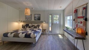 Une chambre avec un lit, une petite table, un banc, un luminaire suspendu et une porte donnant sur l’extérieur.