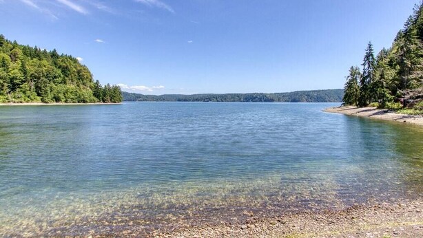 Un lac paisible aux eaux limpides, entouré d’une forêt dense et bordé de collines à l’horizon.