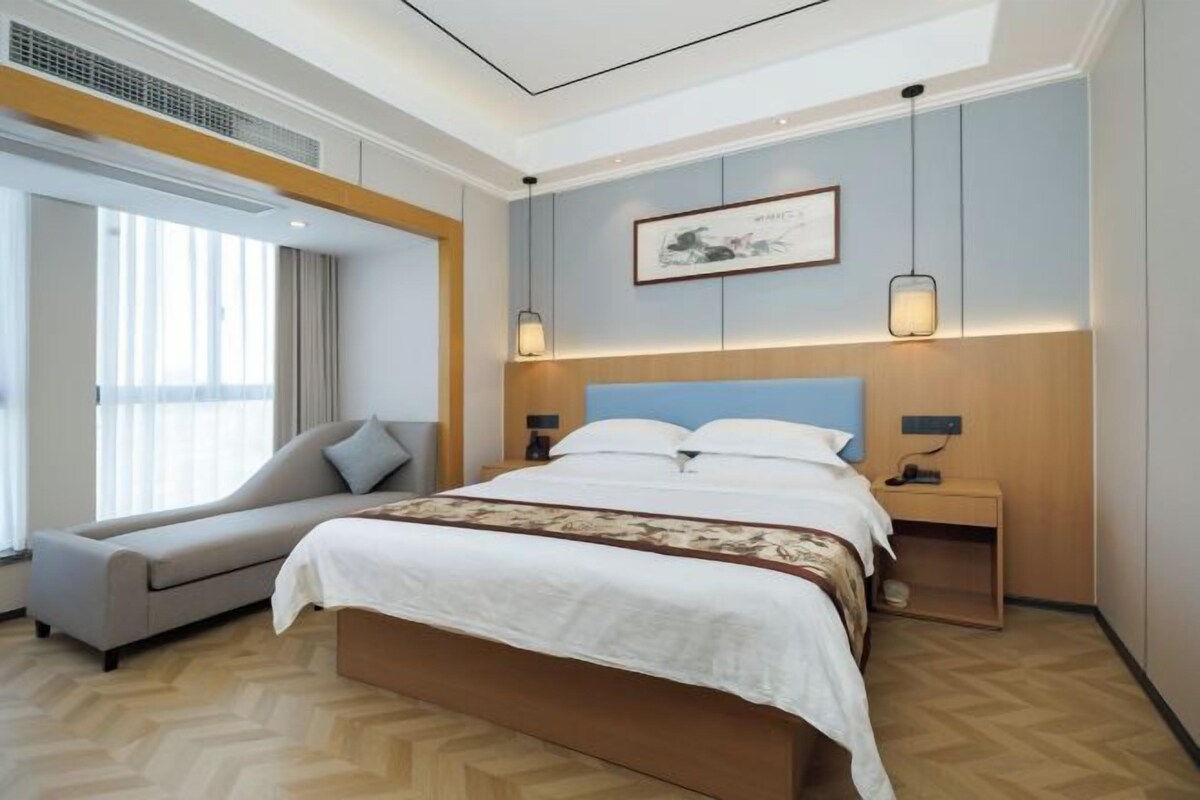 A modern hotel room with a large bed, a sofa, a nightstand, and a wall-mounted artwork.