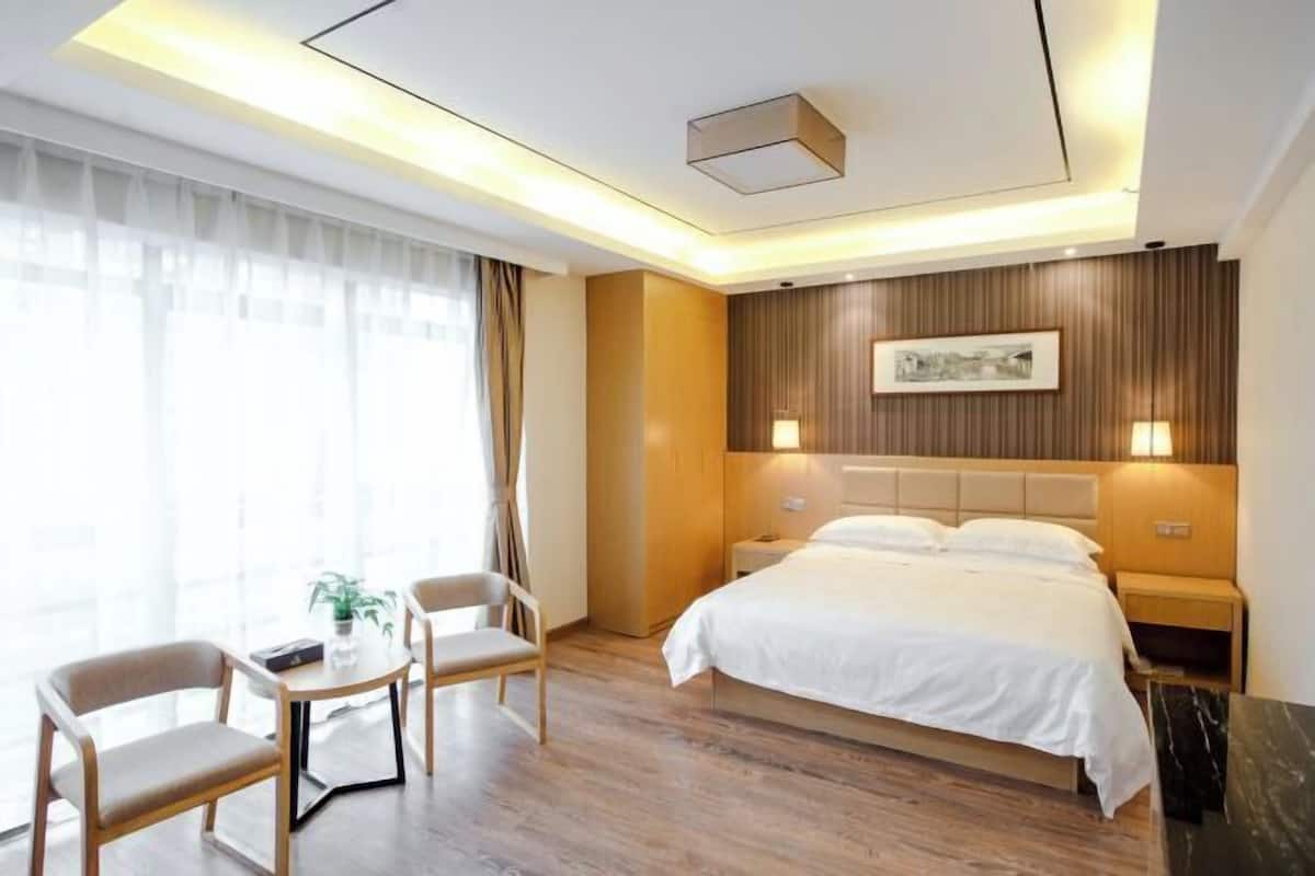 Habitación de hotel con cama, mesitas de noche, un escritorio con sillas, una ventana con cortinas y un cuadro colgado en la pared.