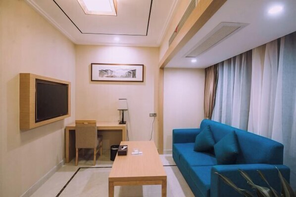 A compact hotel room with a desk, a blue sofa, a television mounted on the wall, and a framed picture above the desk. - Jia Yue Hotel (Tongxiang)