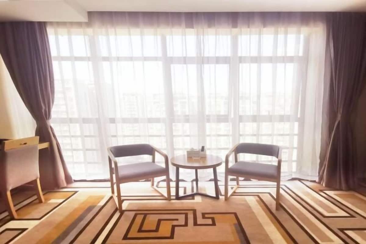 Una habitación de hotel con un ventanal grande, dos sillas, una mesita y una alfombra estampada.