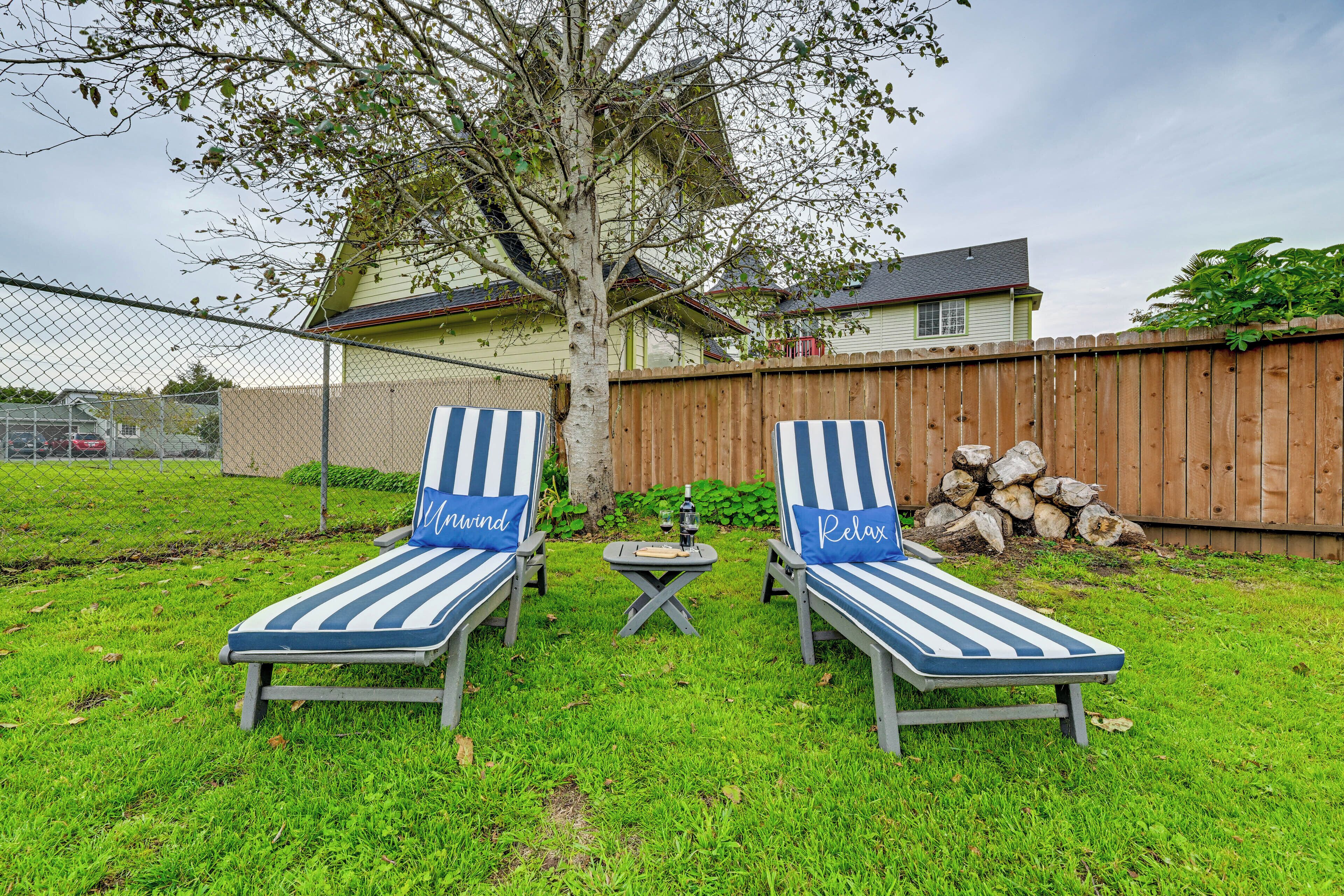 Dos sillones de mimbre a rayas con almohadones que dicen "Mornwind" y "Relax" en un patio con césped, cerca hay una valla de madera y un árbol.