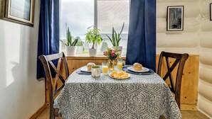 Een eettafel met een tafelkleed met dessin, omringd door houten stoelen, met uitzicht op een raam, potplanten en ontbijtartikelen.