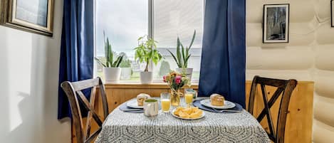 Ett matbord med ett mönstrat bordsduksöverdrag, omgivet av trästolar, med utsikt genom ett fönster, krukväxter och frukostmat.