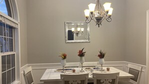Comedor con una mesa blanca, cuatro sillas, una araña y tres jarrones con flores.