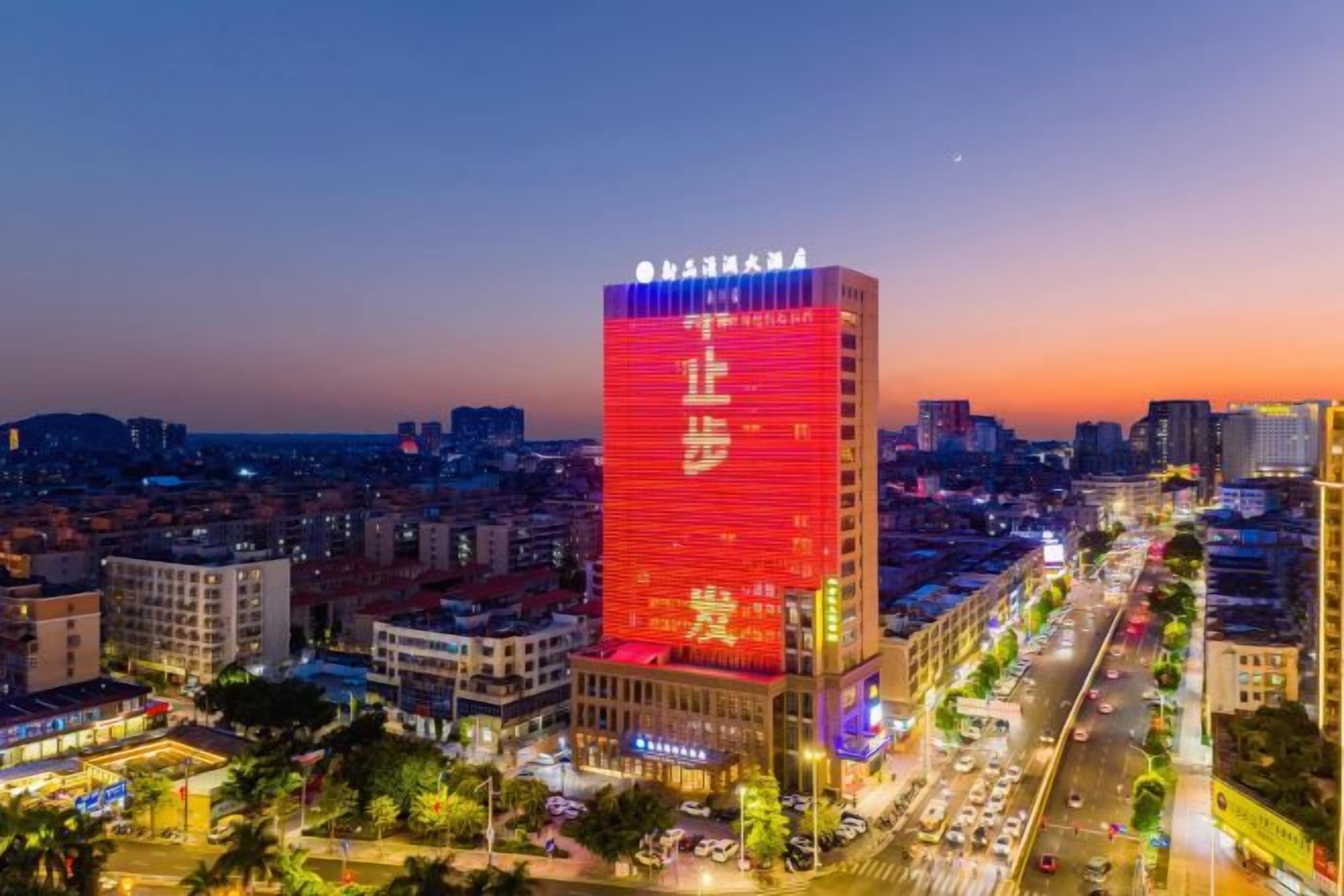 Um edifício alto com letreiros iluminados, cercado por um horizonte urbano ao entardecer.