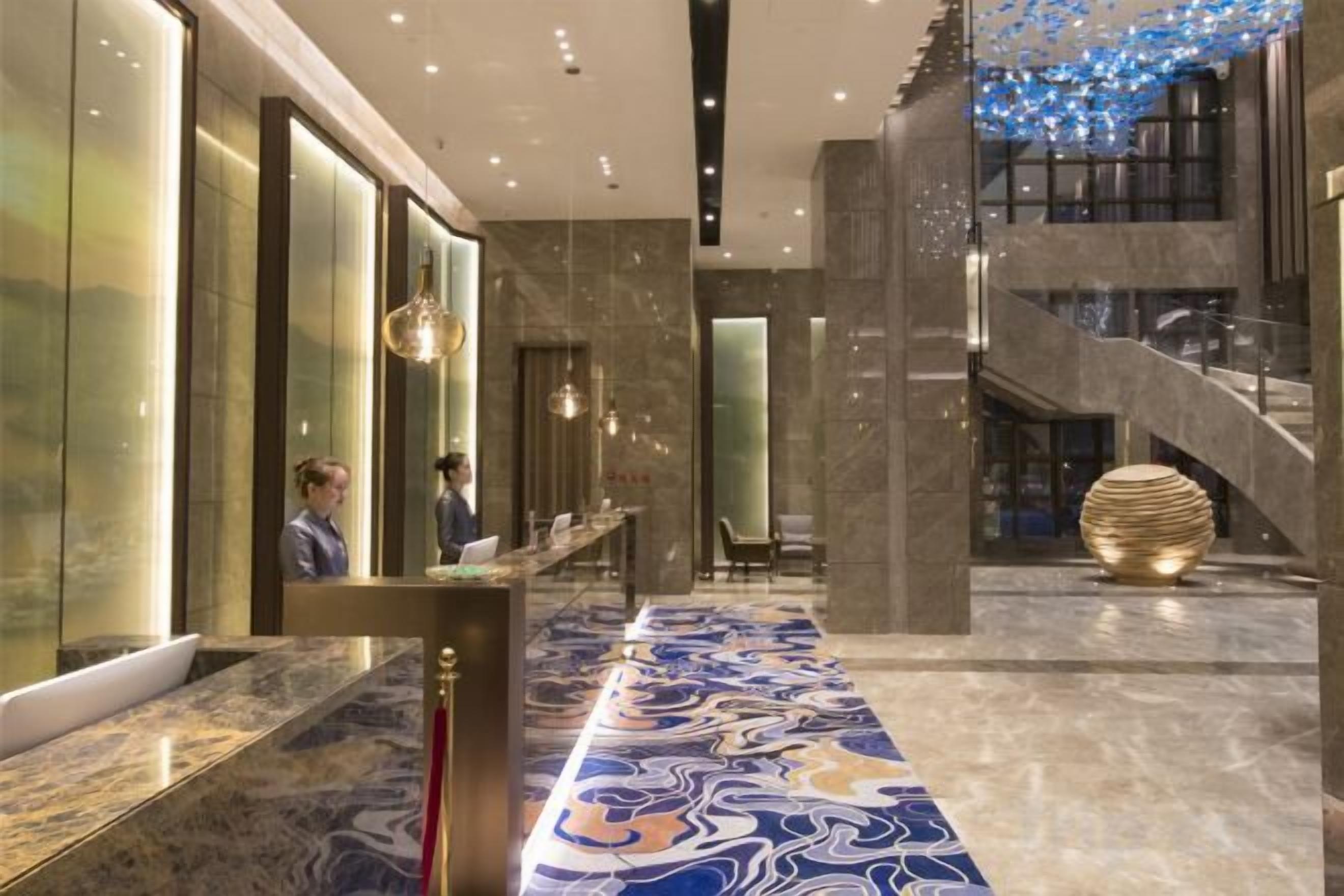A hotel lobby with a marble reception desk, a staircase, and a decorative light fixture.