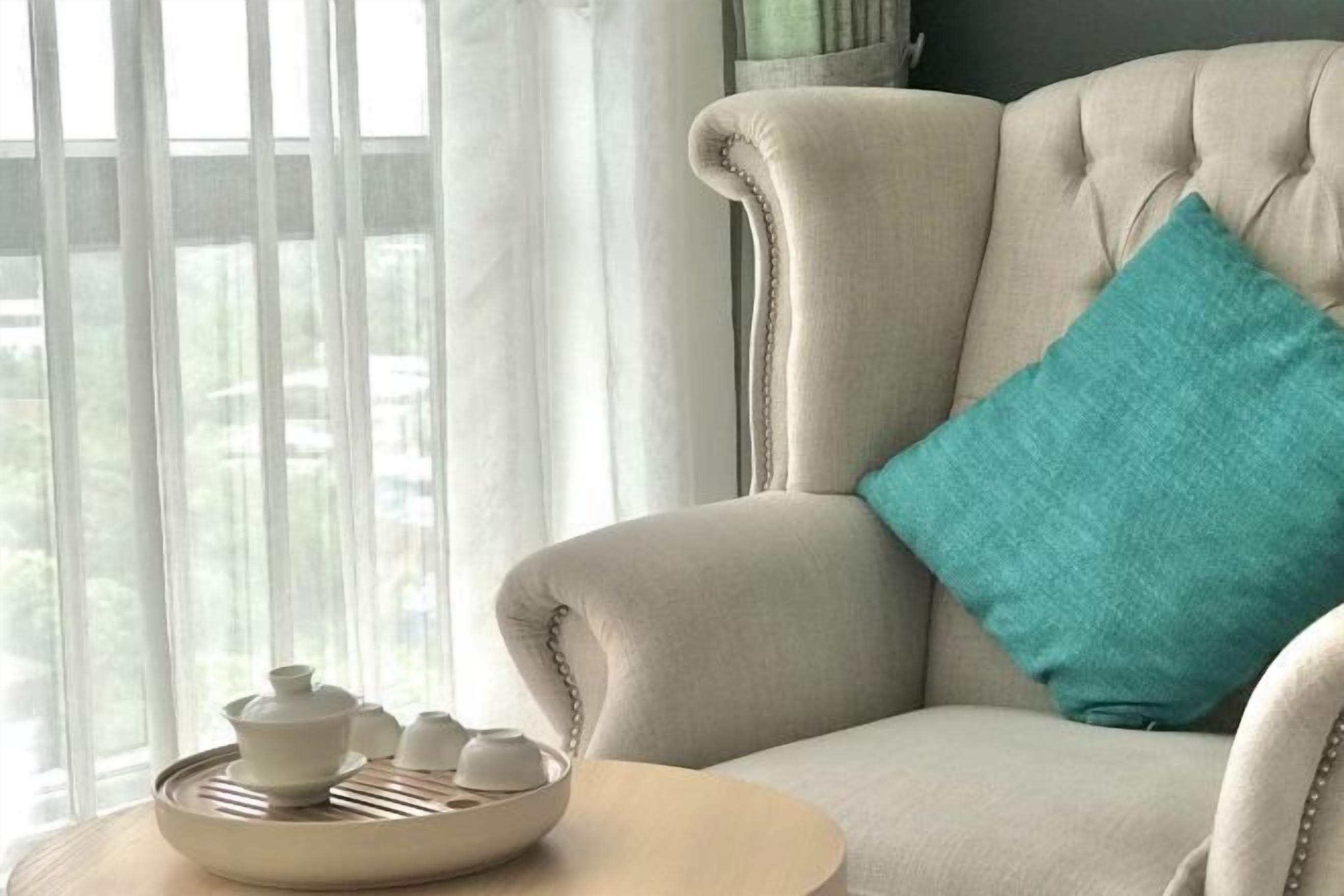 A beige upholstered armchair with a teal pillow, a round table with a tray of teaware, and a window with sheer curtains.