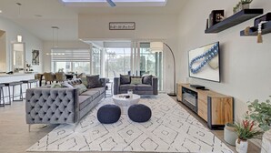 A modern living room with a grey sofa, a coffee table, and two round ottomans. There is a TV mounted on the wall, a ceiling fan, and a large window providing natural light. - Blue Pearl| Pool | Hot Tub | 3 Bed, 2 Bath (Rotonda West)