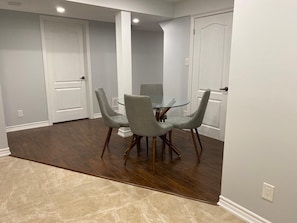 Uma área de refeições com uma mesa de vidro e quatro cadeiras cinzentas, piso de madeira e paredes brancas.