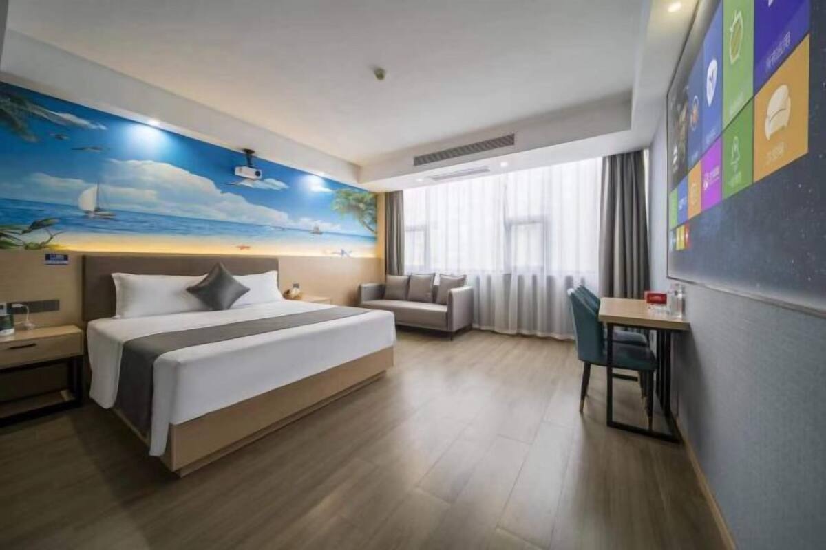Une chambre d’hôtel comprenant un lit, un bureau, une chaise, un canapé, une fresque représentant une scène de plage et une fenêtre avec des rideaux.