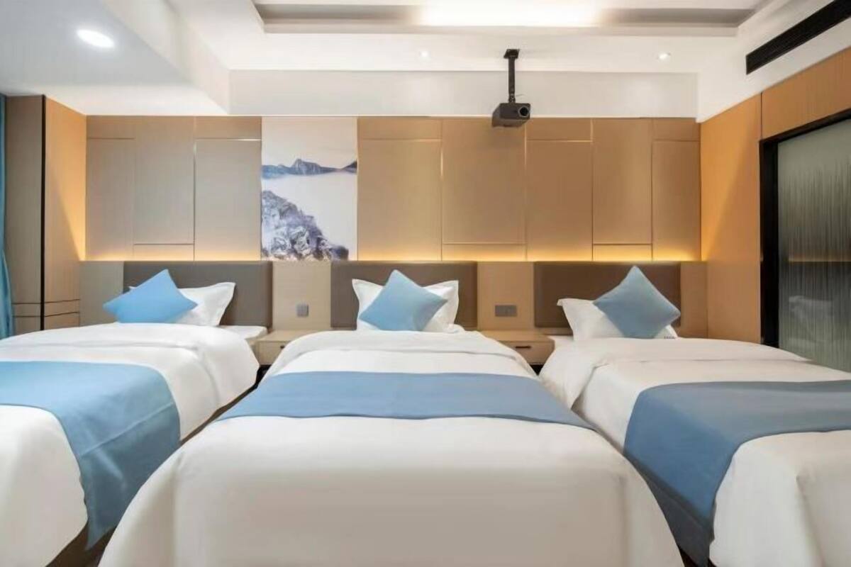 Une chambre d’hôtel avec trois lits, chacun recouvert d’une couverture bleue et blanche. La chambre est équipée d’un grand téléviseur fixé au mur et d’un projecteur au plafond.