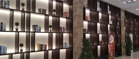 A modern hotel lobby with a long reception desk, a marble floor, and a wall of bookshelves with potted plants.