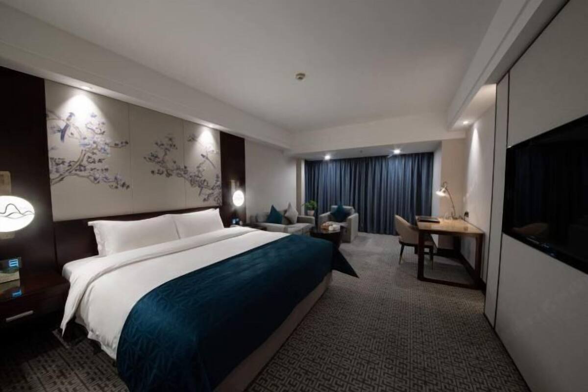 Une chambre d’hôtel avec un lit, un bureau, une chaise, une lampe et une grande fenêtre avec des rideaux.
