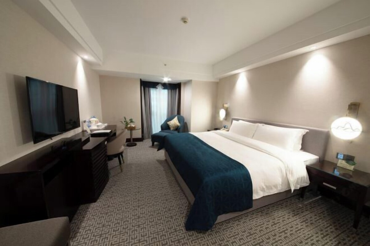 A hotel room with a large bed, a desk with a chair, a television, and a bedside table with a lamp.