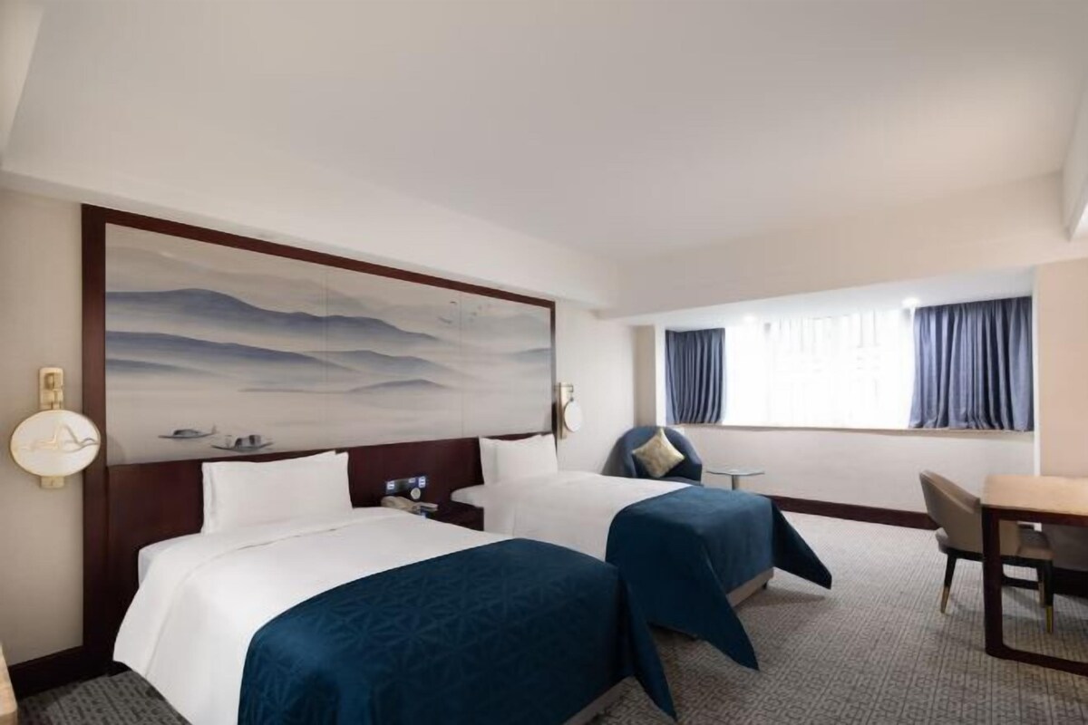 A hotel room with two beds, a desk, a chair, a large abstract painting on the wall, and a window with curtains.